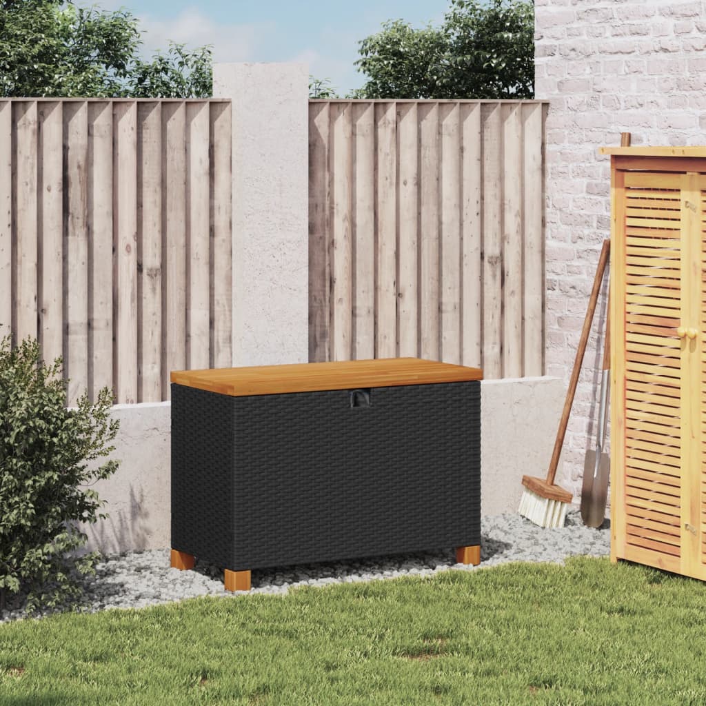 Zwarte rattan opbergbox voor buiten met acaciahouten deksel en poten, geplaatst op een groen gazon tegen een houten hek achtergrond.